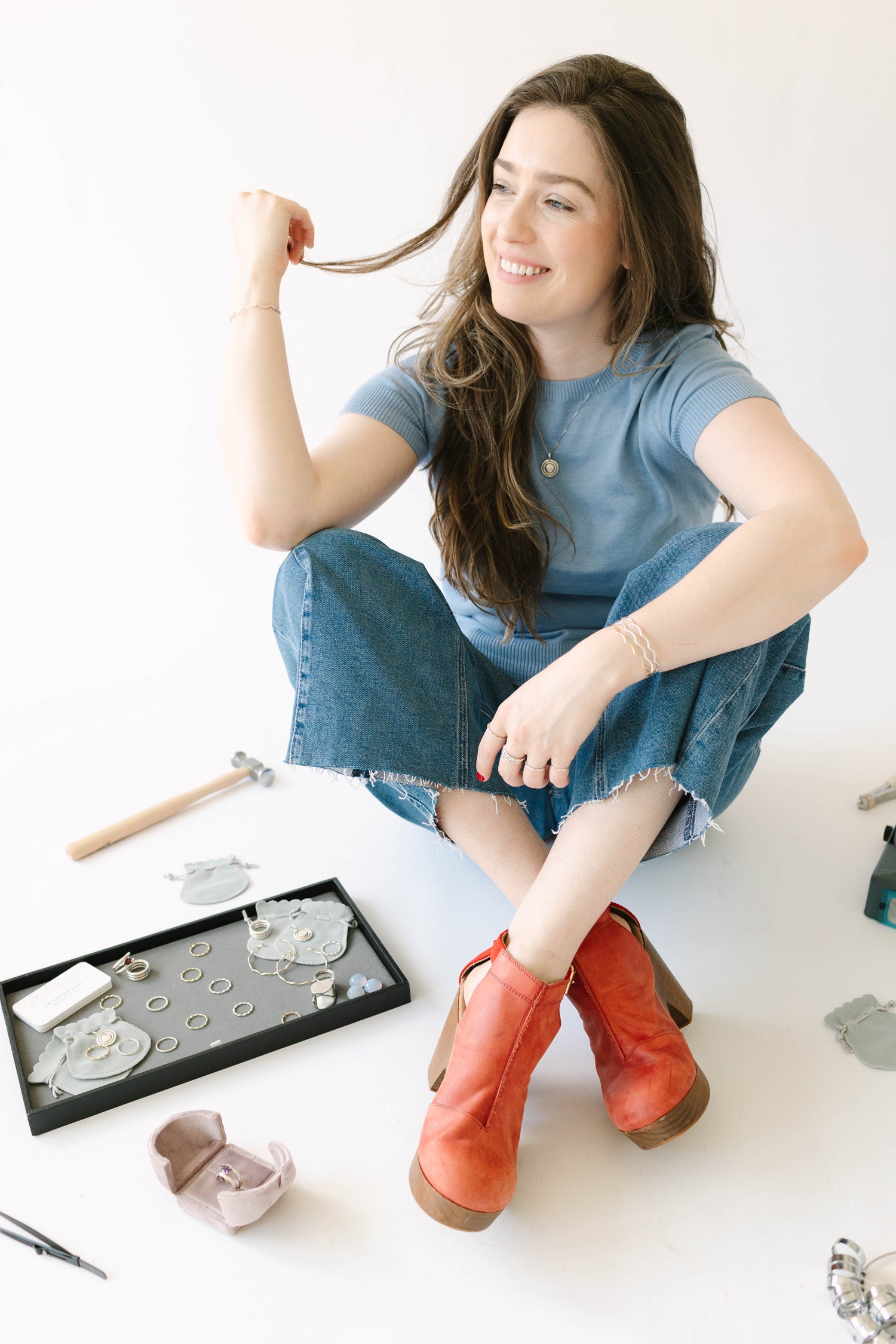 Jewelry designer Lily Wujek sits on the floor with white background, wearing red shoes and jewelry from her collection and surrounded by her jewelry-making tools